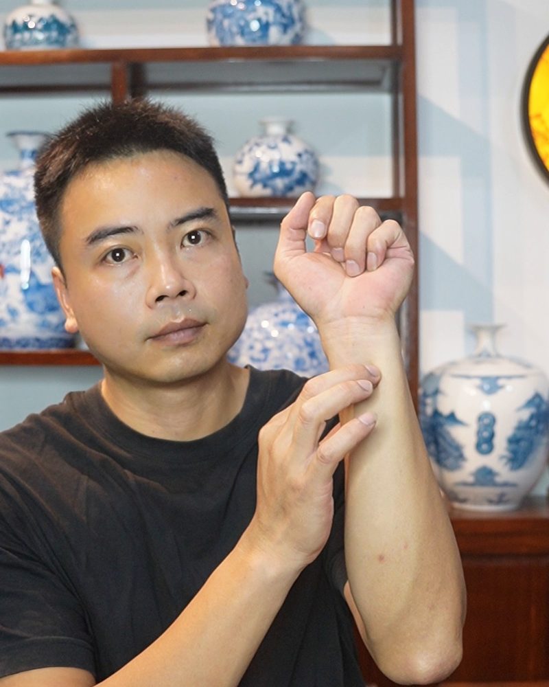 Nguyen Duy Phong pressing the inside of his wrist with two fingers, in front of a shelf of blue-and-white vases